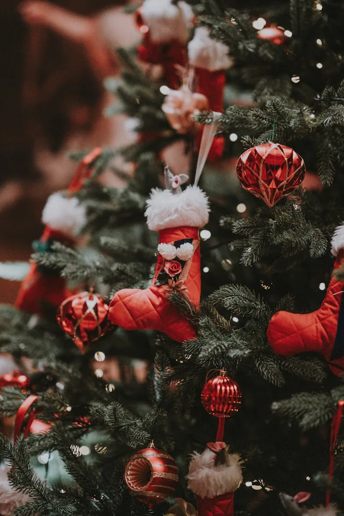 Santa Claus stands proudly beside a richly decorated Christmas tree with sparkling lights, gold and red ornaments, and evergreen branches. The warm glow from the tree lights illuminates his red suit and white beard.