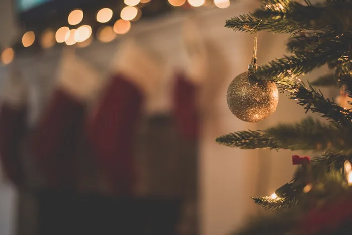 Close-up of a white brick fireplace mantel decorated for Christmas with fresh evergreen garlands, red berries, candy canes, and three hanging stockings with white fur trim. Small holiday gnome figurines sit scattered across the hearth below.