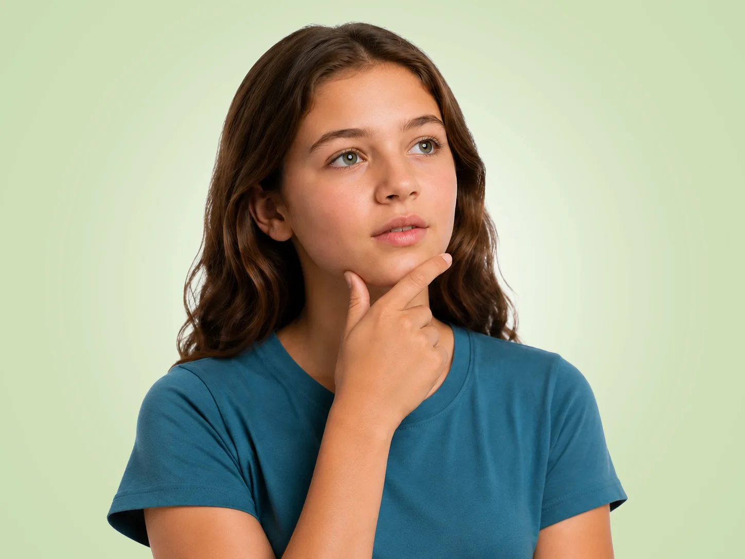 A young girl with wavy brown hair in a teal t-shirt, looking up thoughtfully with her hand on her chin.