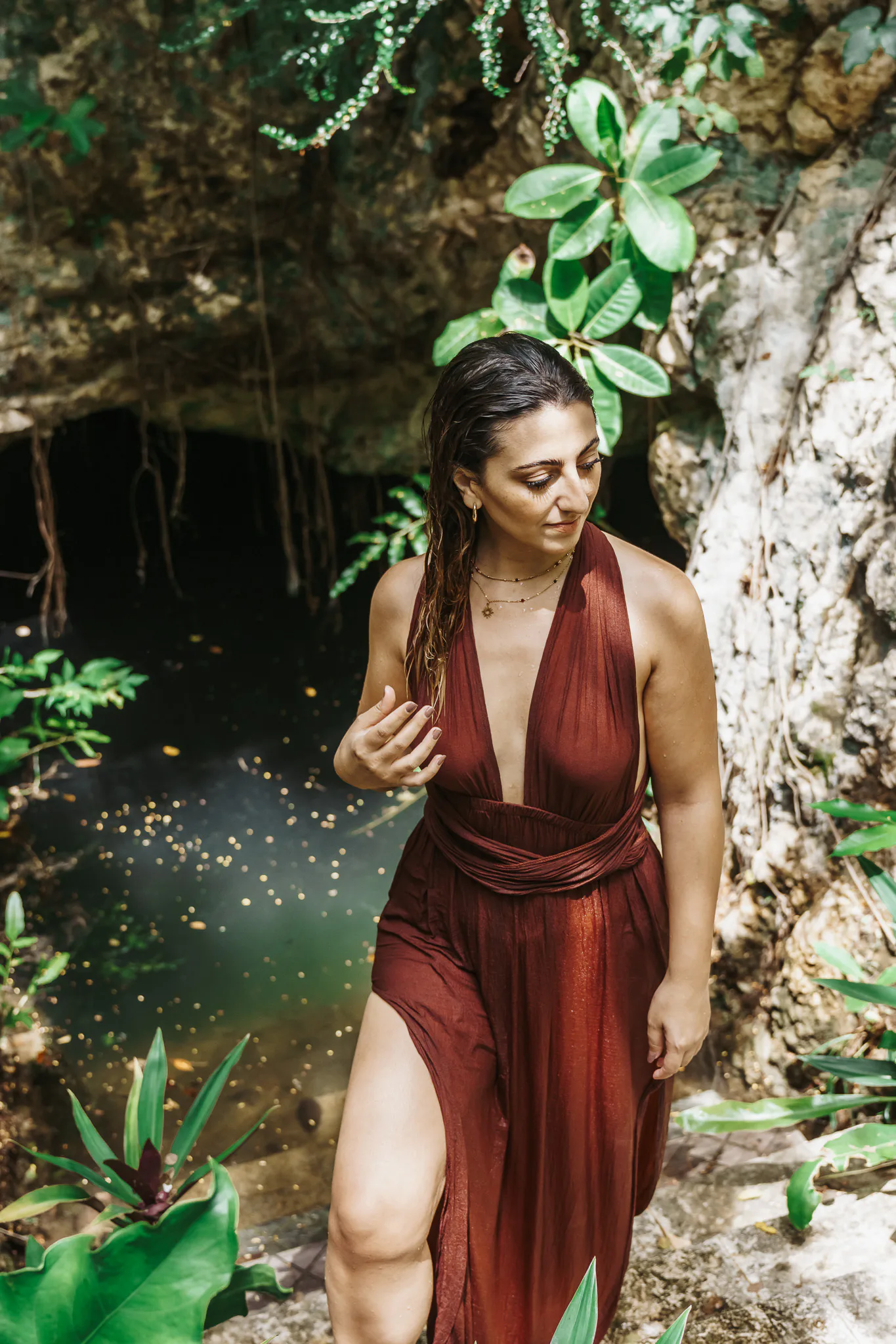 Ivonne Zarur de pie junto a un cenote, con vestido largo color vino, mirada baja y serena, luz natural cálida.